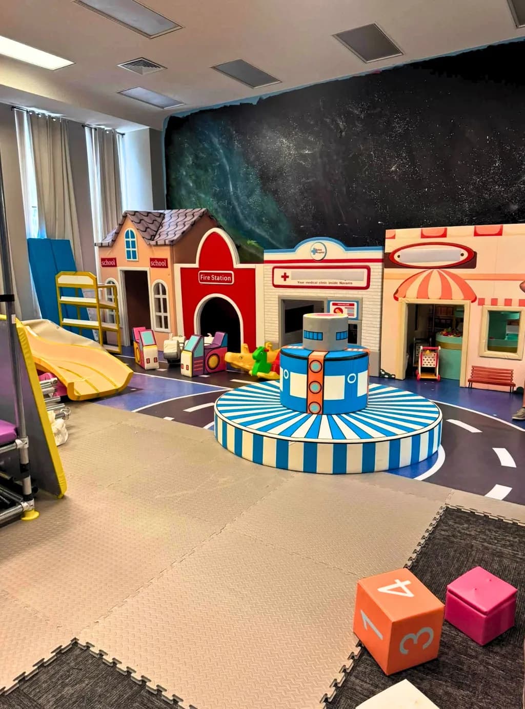 Kid sized town with school, fire station, and clinic fronts around a striped carousel.