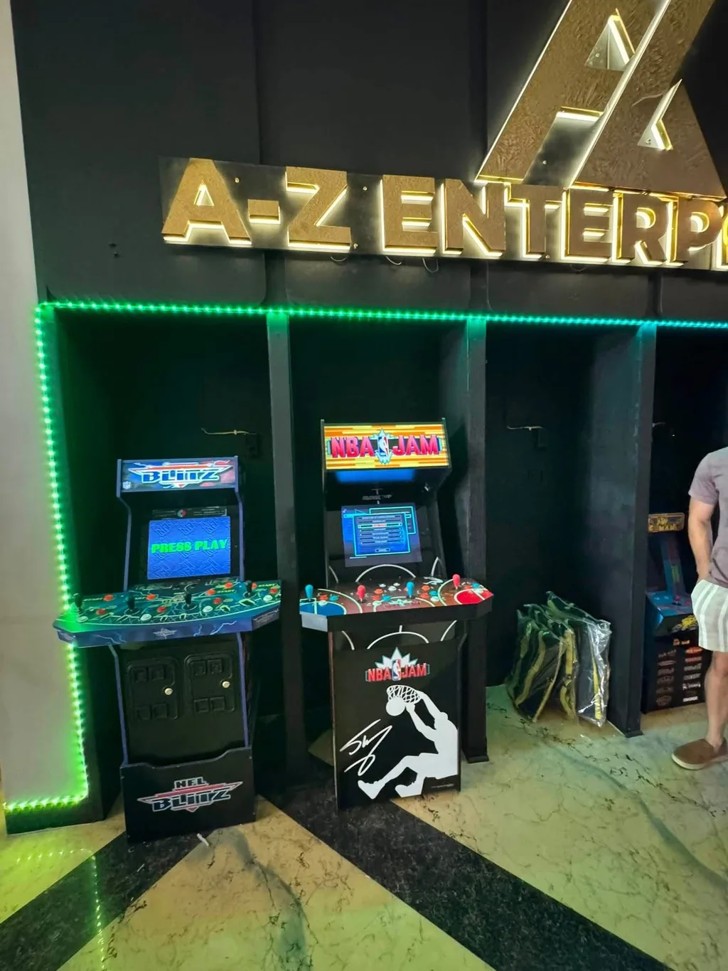 Two arcade cabinets, NFL Blitz and NBA Jam, lit by green LED under the gold A-Z Enterprises sign.